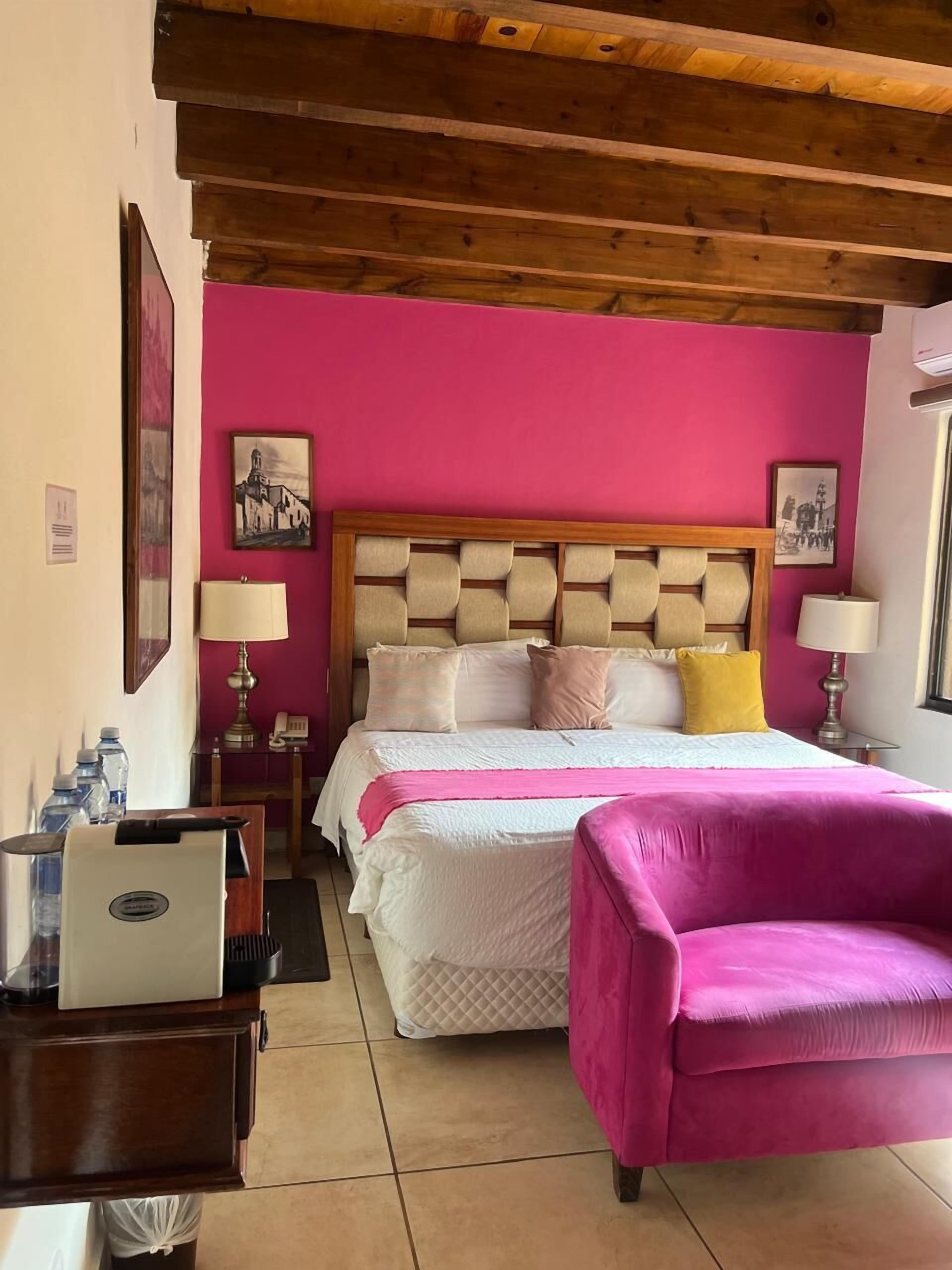 A hotel room with a bed, a desk with a laptop, a pink armchair, and a wooden ceiling.