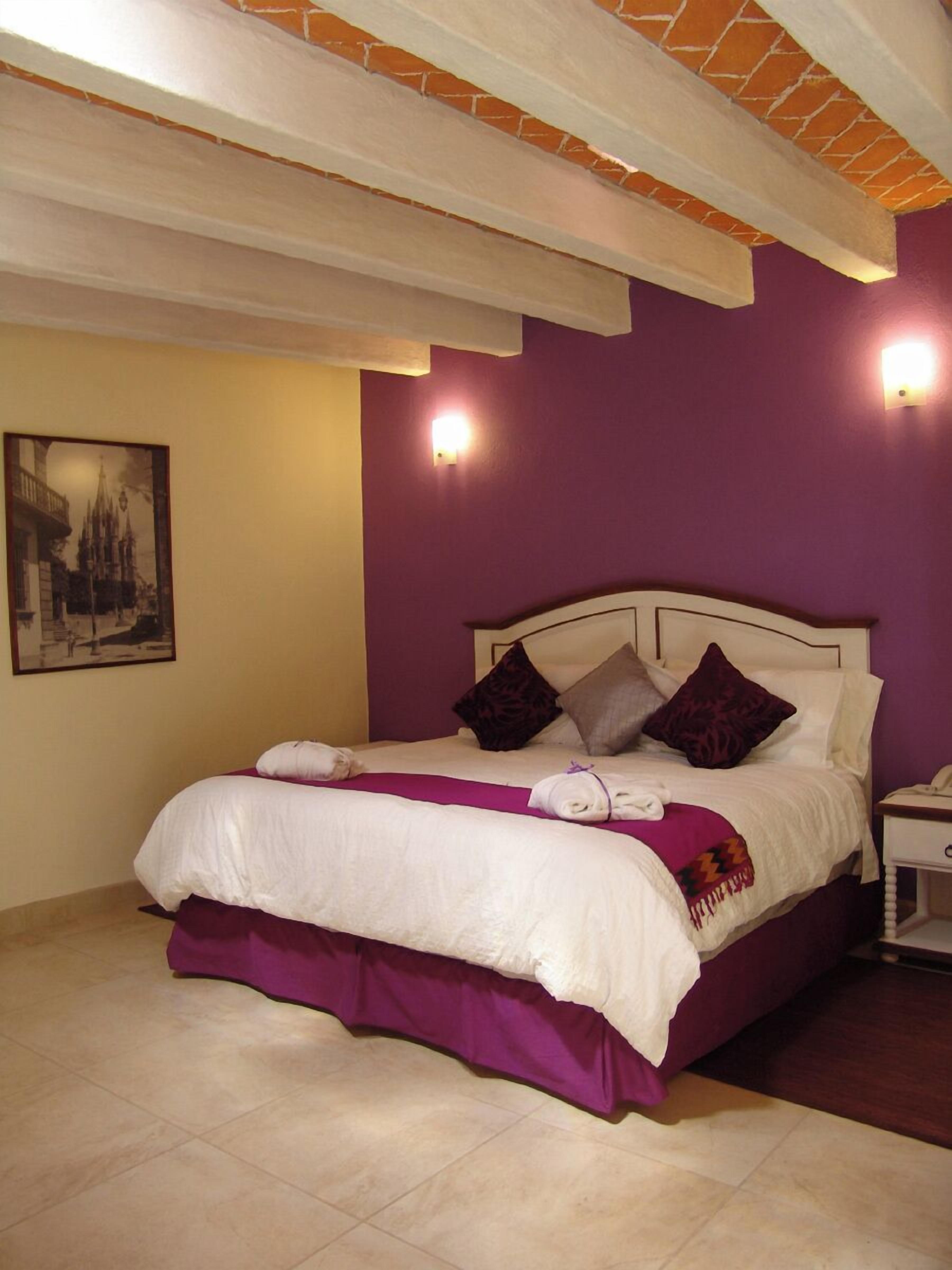 A neatly made bed with a purple bed skirt and white linens, a nightstand with a lamp, and a framed picture on the wall.