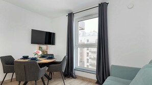 Een moderne woonkamer met een eethoek, een flatscreen televisie aan de muur en een groot raam met gordijnen.