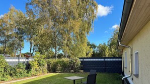 Une terrasse avec une table et des chaises, une plateforme en bois et un jardin agrémenté d’une clôture et d’arbres.