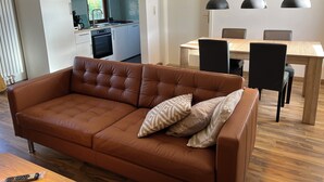 Un salon moderne avec un canapé en cuir marron, une table à manger en bois avec des chaises, une cuisine en arrière-plan et un ventilateur fixé au mur.