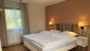 Un lit double avec une literie blanche, une tête de lit en bois, deux tables de chevet avec des lampes, une fenêtre avec des rideaux et un plancher en bois.
