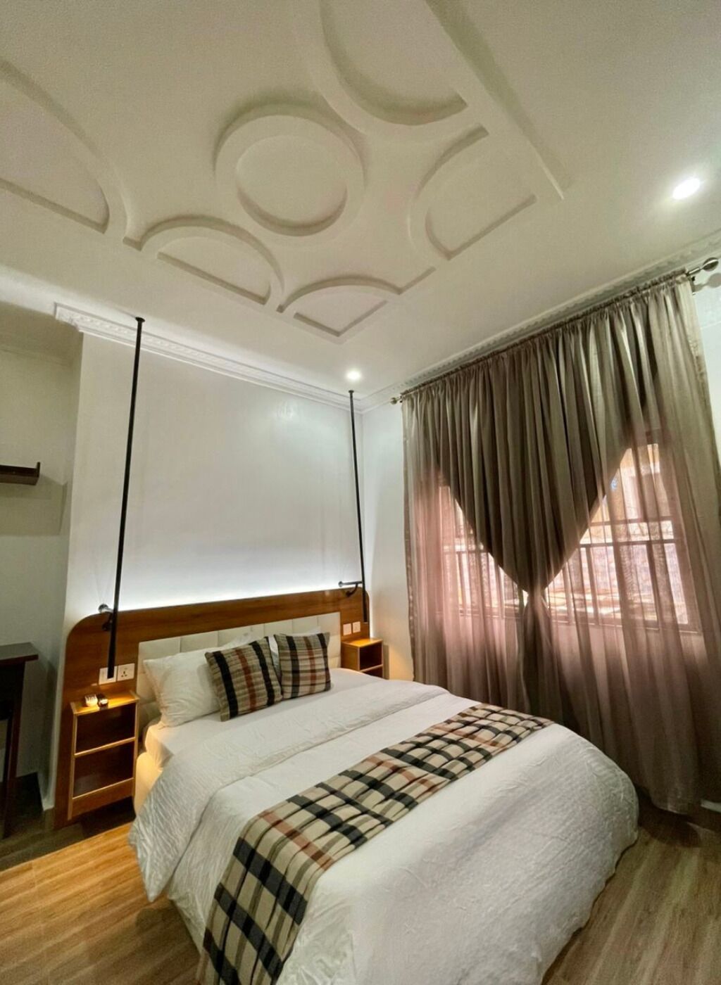 Une chambre à coucher avec une tête de lit en bois, un grand lit recouvert d’une couverture à carreaux, une table de chevet et une fenêtre avec des rideaux.