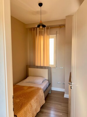 Una habitación pequeña con cama individual, piso de madera, una colcha texturizada, una lámpara colgante y una ventana con cortinas.