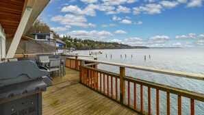 Une terrasse en bois avec des sièges d’extérieur et un barbecue, offrant une vue sur un lac paisible dont la rive s’étend au loin.