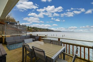 Une terrasse extérieure avec une table et des chaises en bois, offrant une vue sur une étendue d’eau et une côte lointaine.
