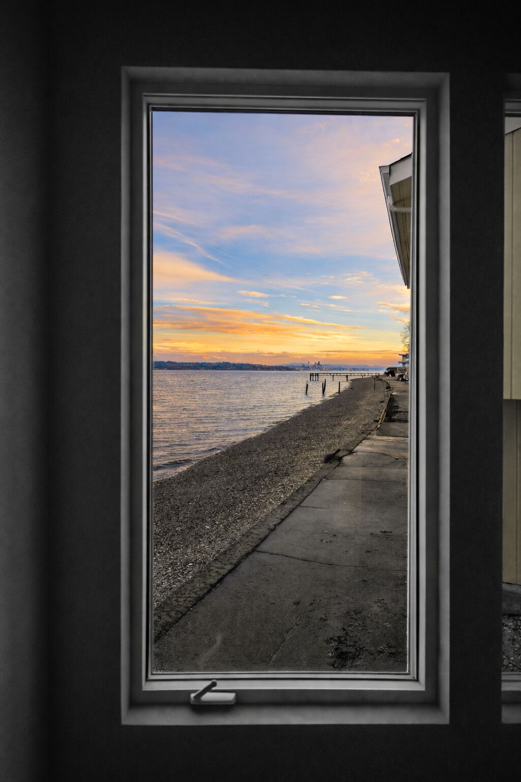 A window with a view of a pebbled beach and a sunset over the water.