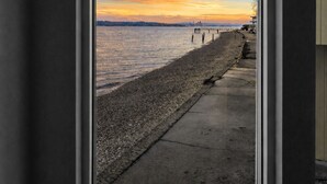 Une fenêtre donnant sur une plage de galets et sur un coucher de soleil sur l’eau.