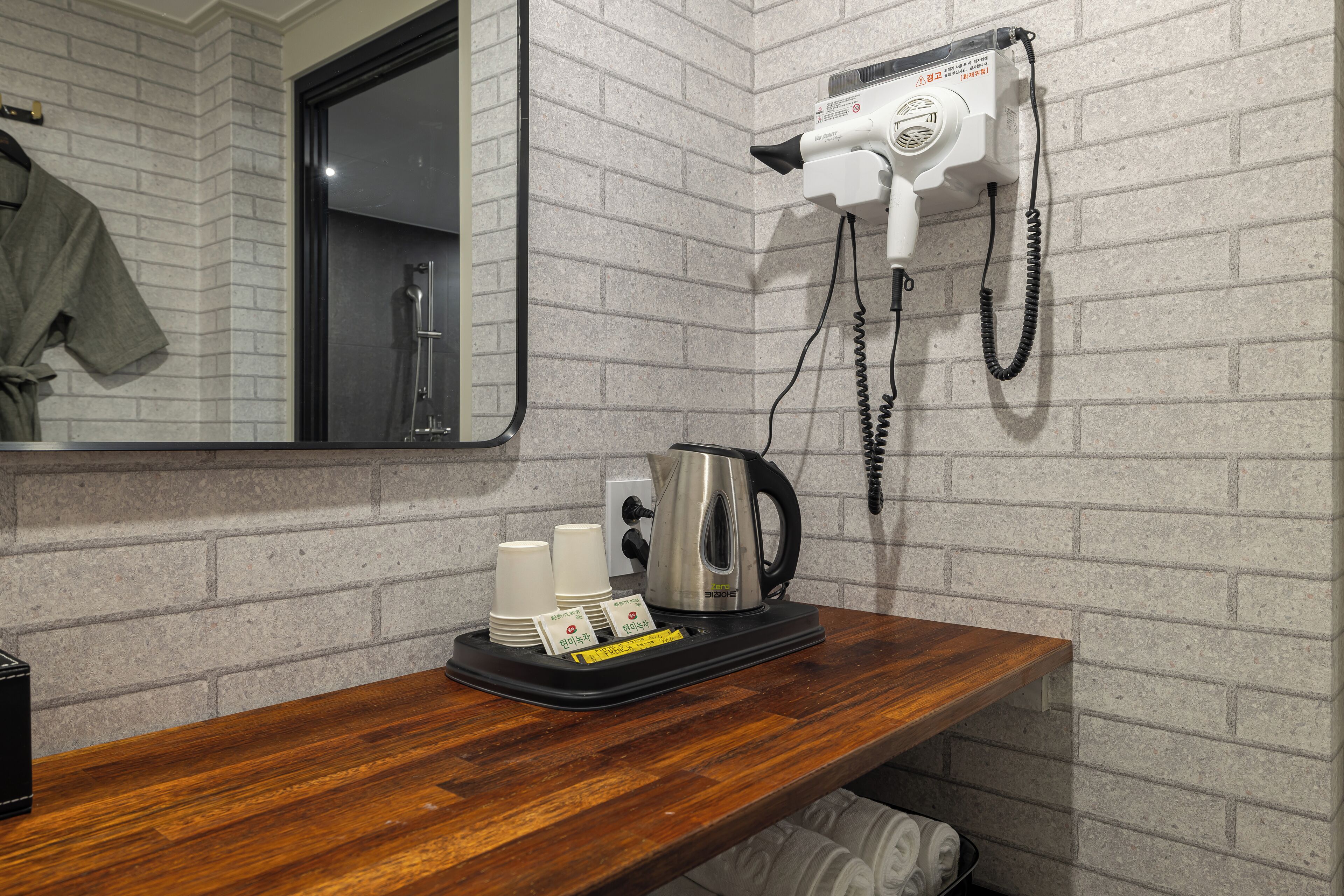 Une salle de bain d’hôtel avec un mur en briques, un meuble-lavabo en bois, un sèche-cheveux fixé au mur, une bouilloire et un porte-serviettes.