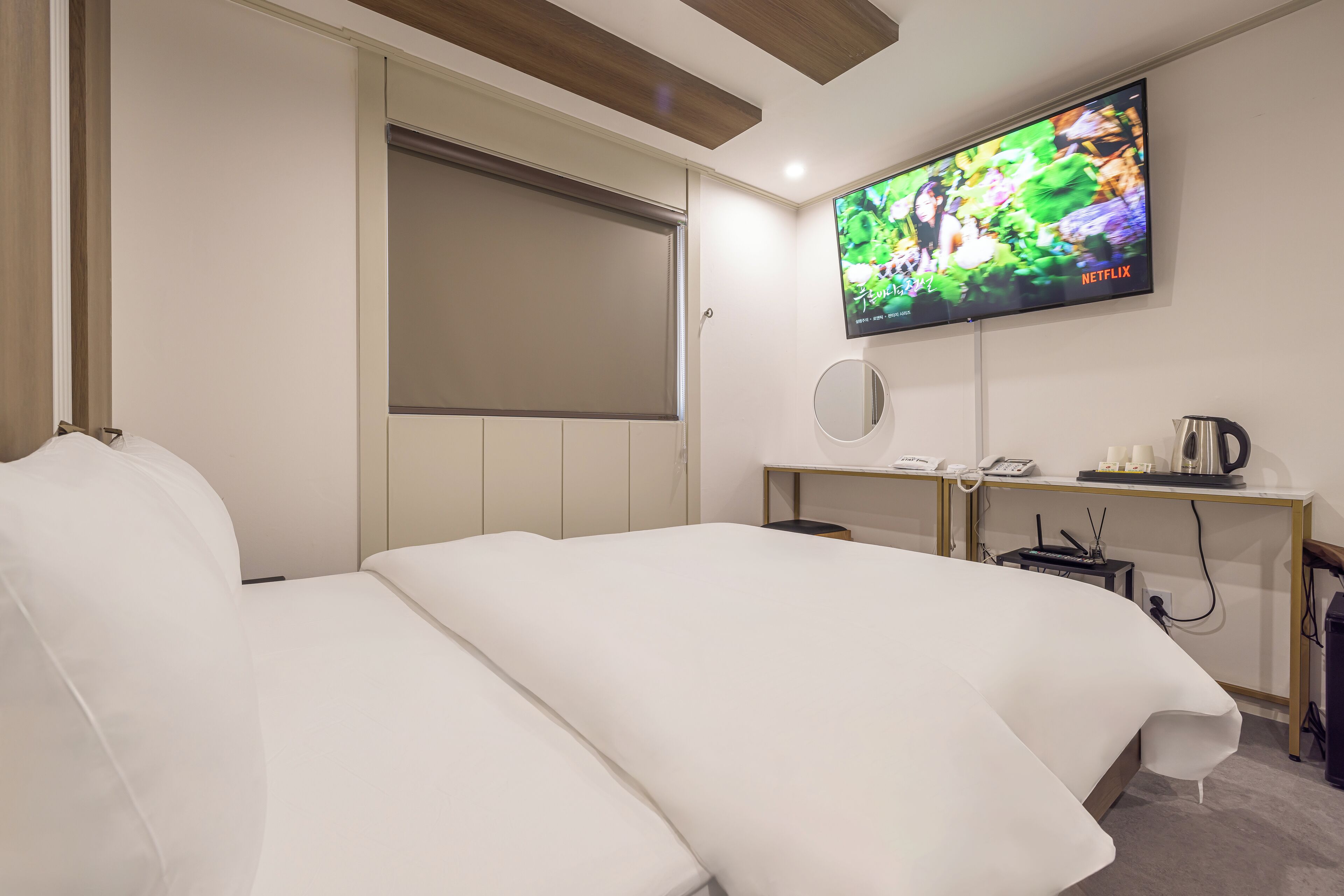 A modern hotel room with a large bed, a flat-screen TV mounted on the wall, a desk with a telephone and a kettle, and a small round mirror.