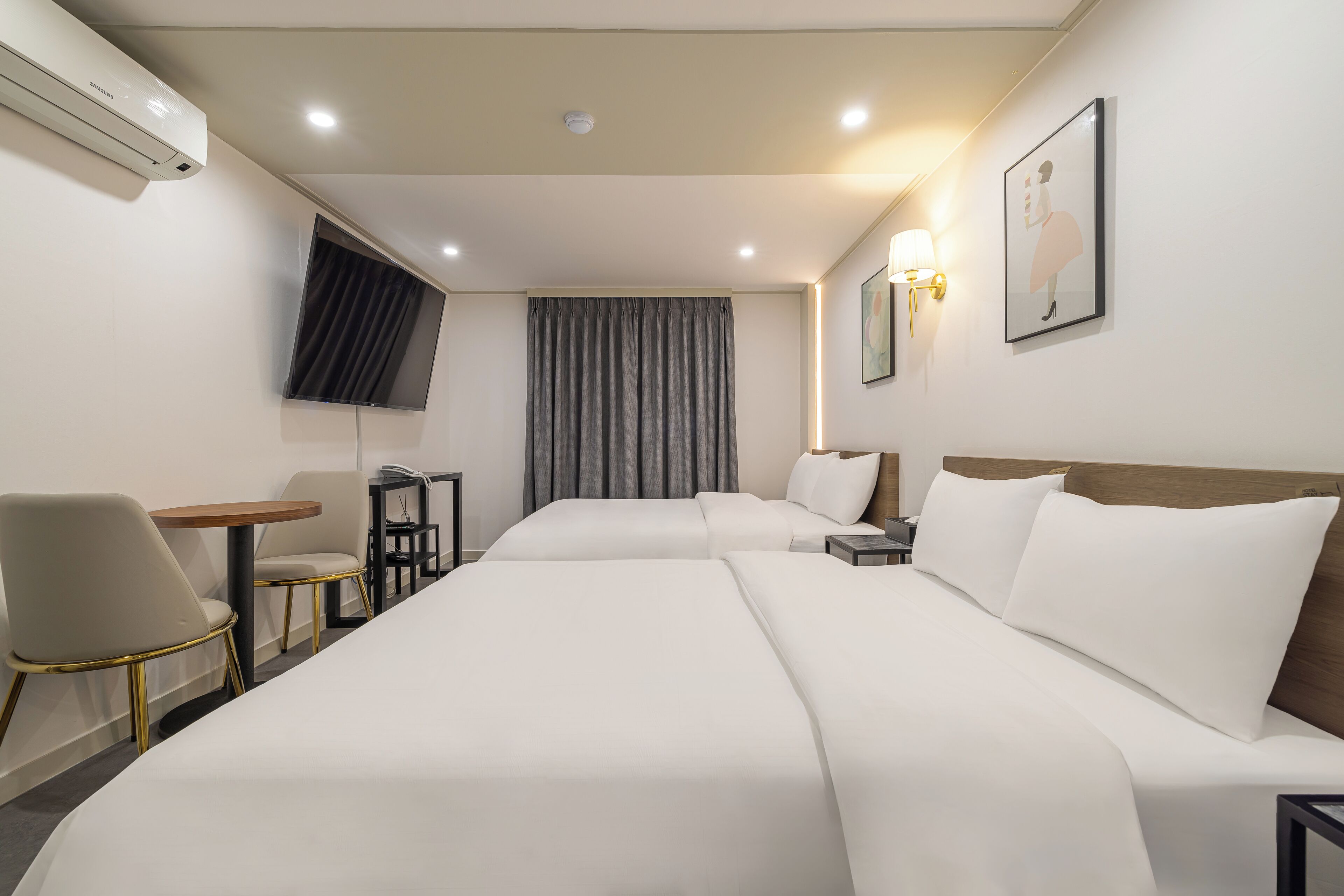 A hotel room with two beds, a desk, a chair, a TV, and a wall-mounted air conditioner.