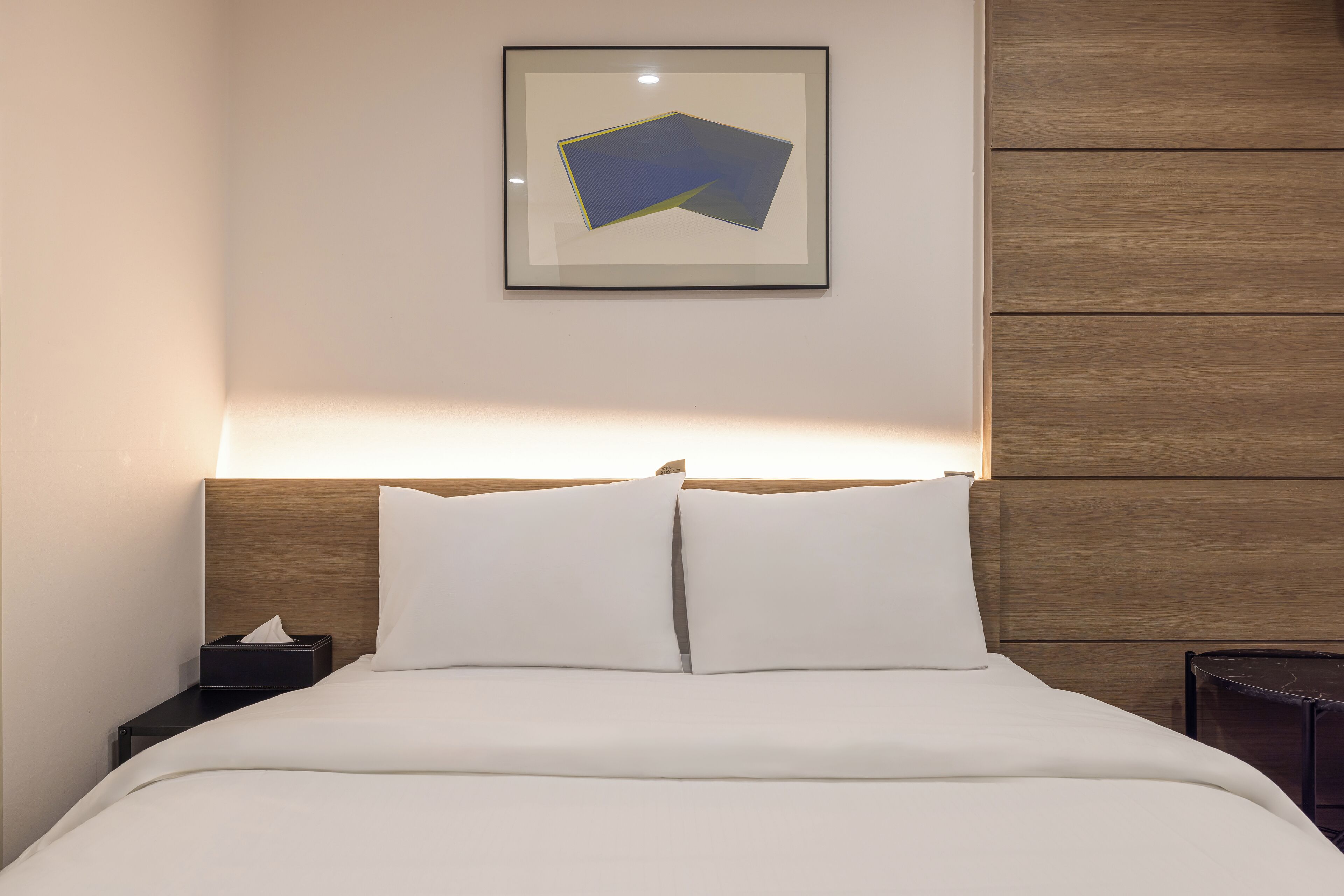 Una cama bien hecha con sábanas blancas, un cabecero de madera y una mesita de noche con una caja de pañuelos.