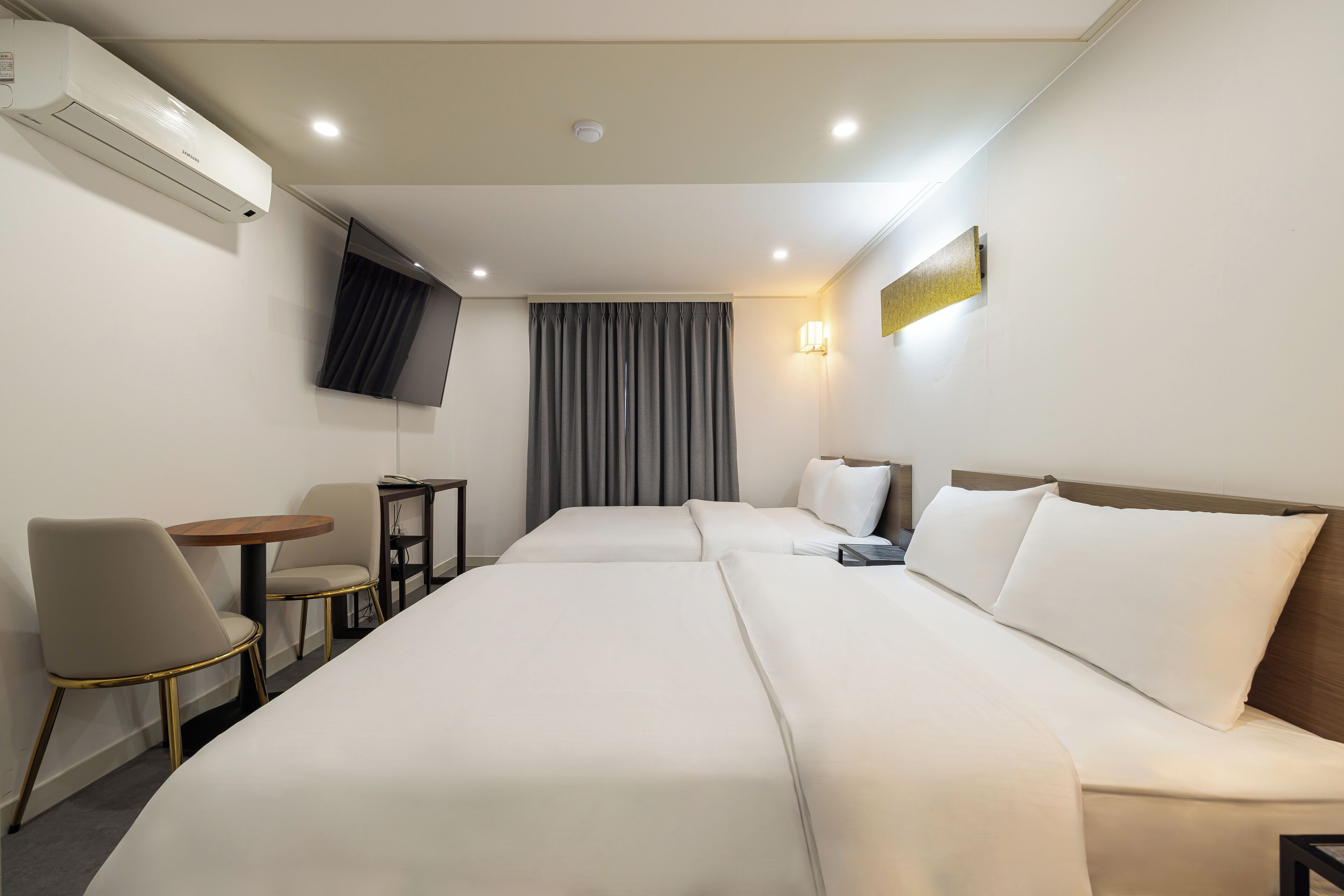 Habitación de hotel con dos camas, un televisor montado en la pared, una mesita con sillas y un aplique de pared.