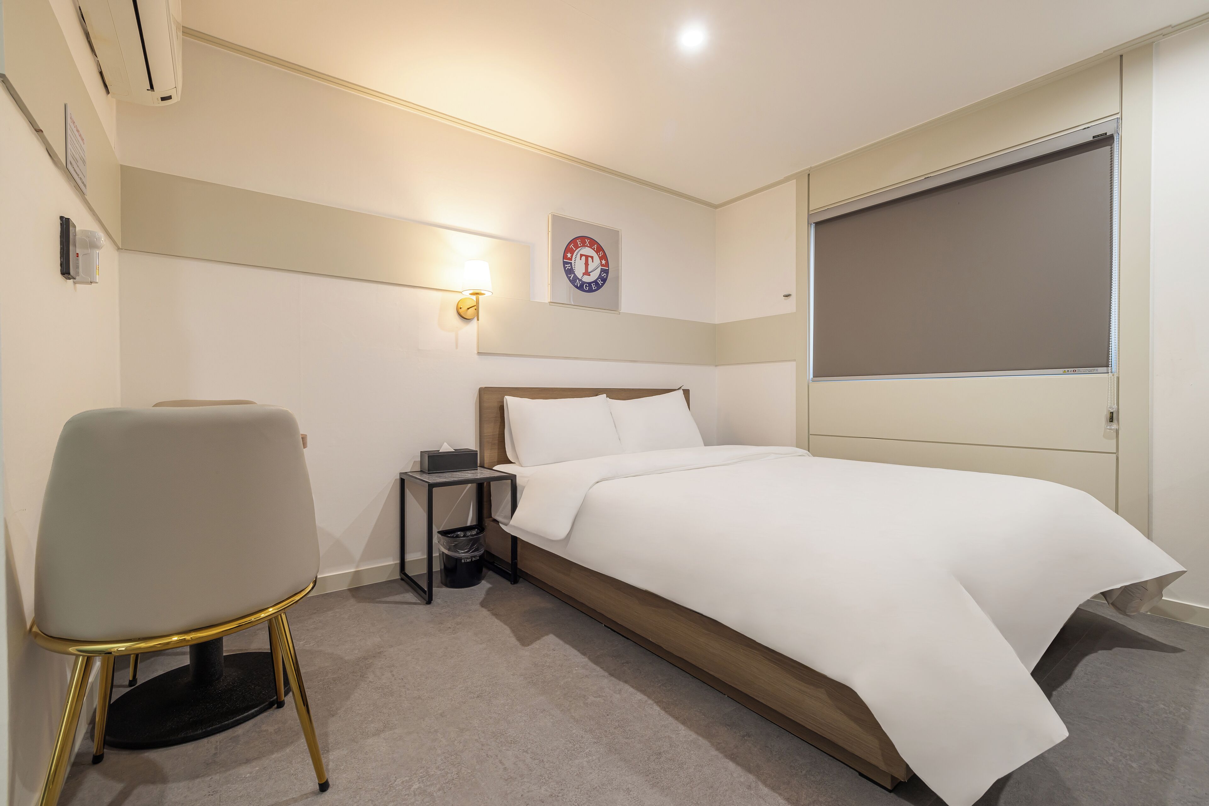 A hotel room with a bed, a chair, a nightstand, a wall-mounted lamp, and a window with a blind.