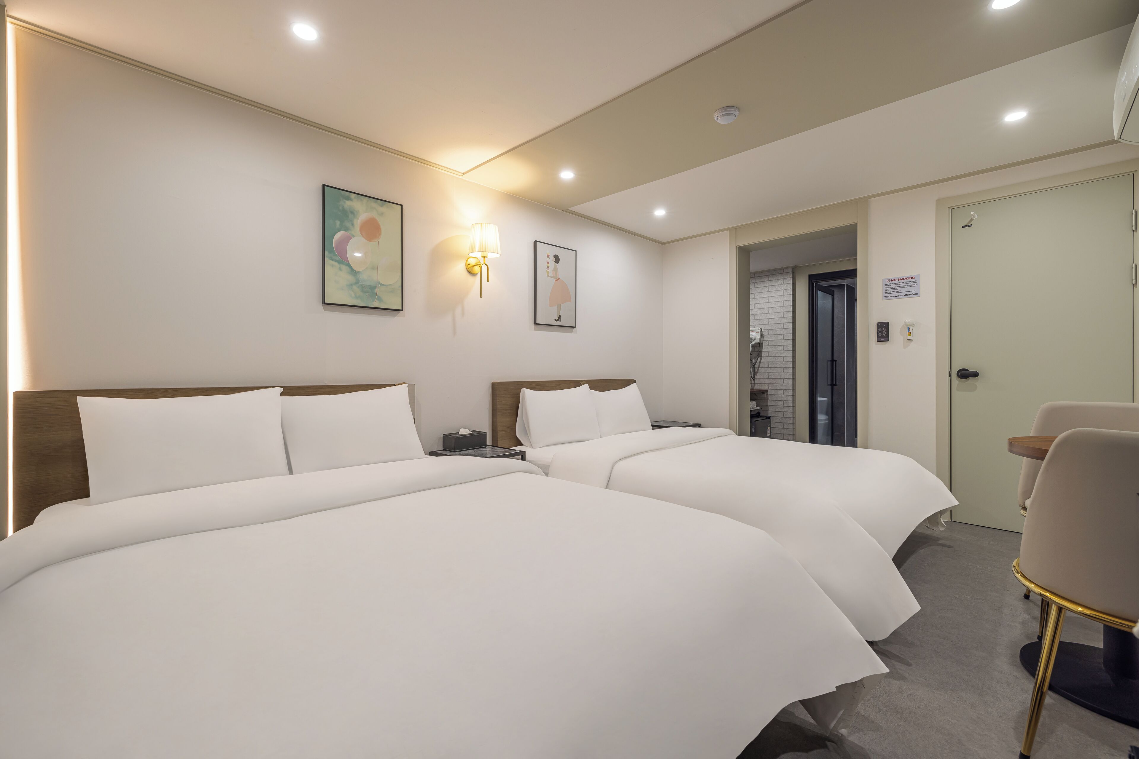 Une chambre d’hôtel avec deux lits, un bureau et une chaise. On y trouve des lampes fixées au mur et une porte avec une pancarte.
