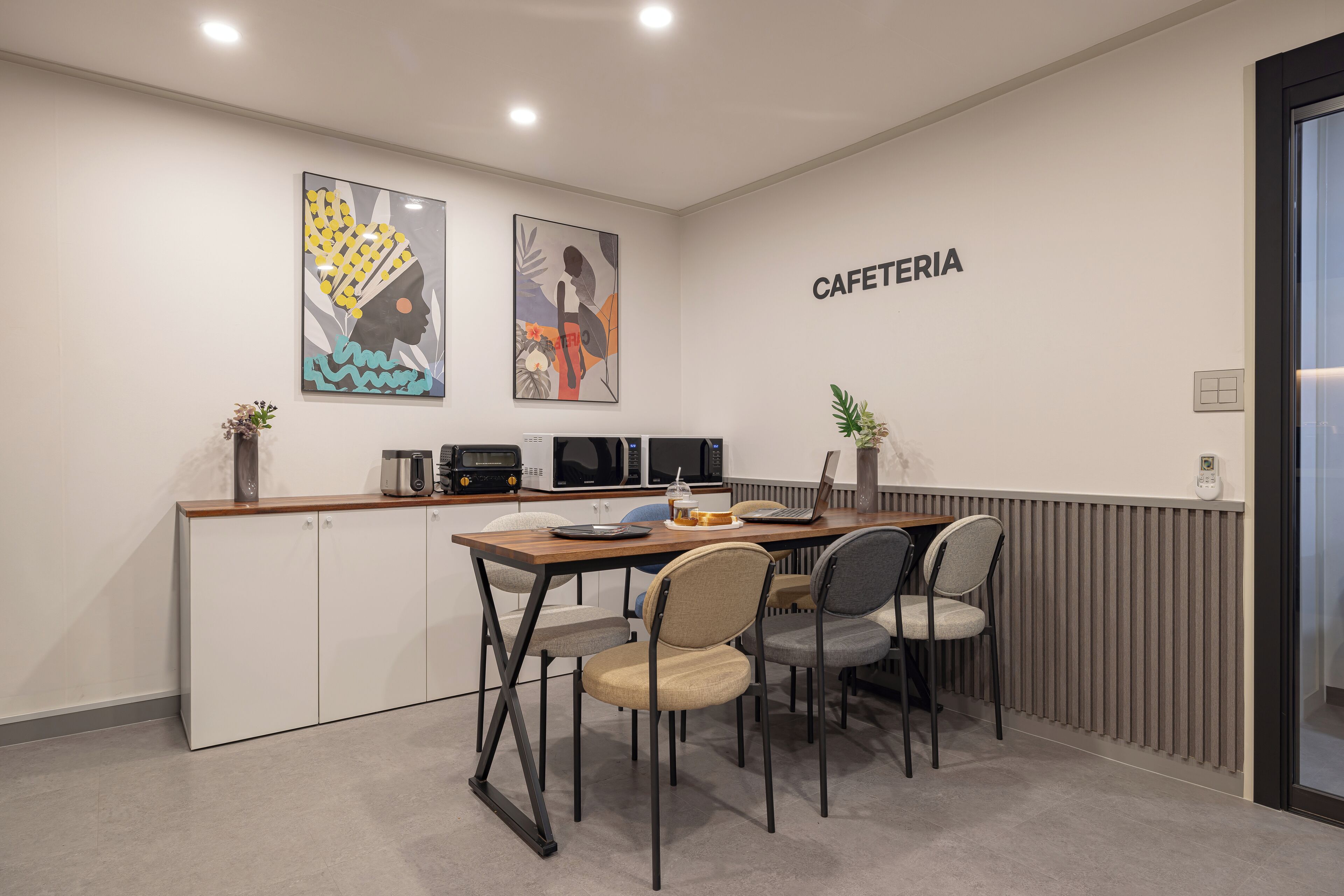 A modern kitchenette with a microwave, toaster, and coffee maker. There is a dining area with a table and chairs, and a sign that reads "CAFETERIA".