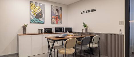 A modern kitchenette with a microwave, toaster, and coffee maker. There is a dining area with a table and chairs, and a sign that reads "CAFETERIA".