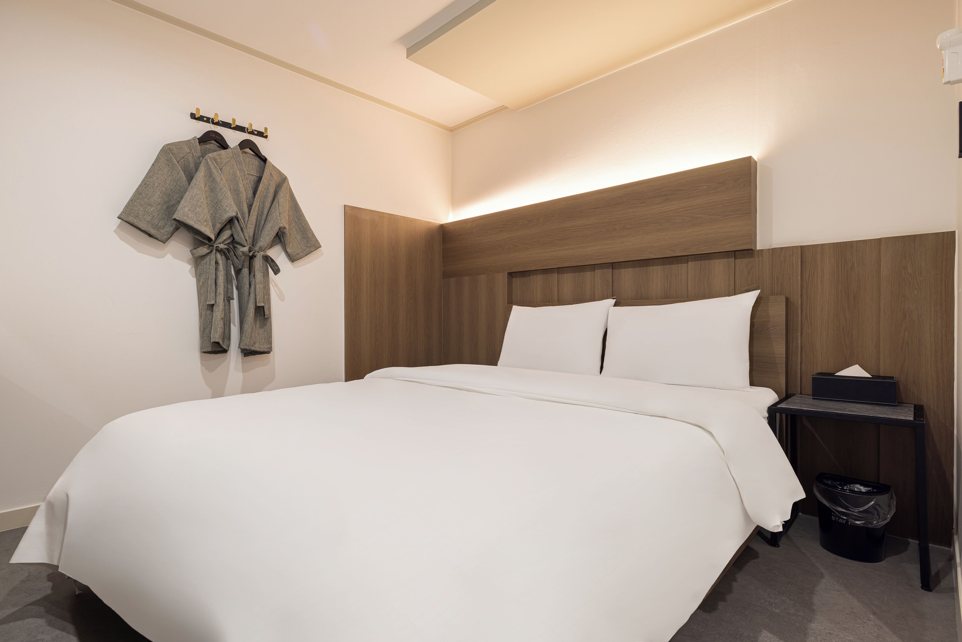 A neatly made bed with white linens, a wooden headboard, and a bedside table with a tissue box and a trash bin.
