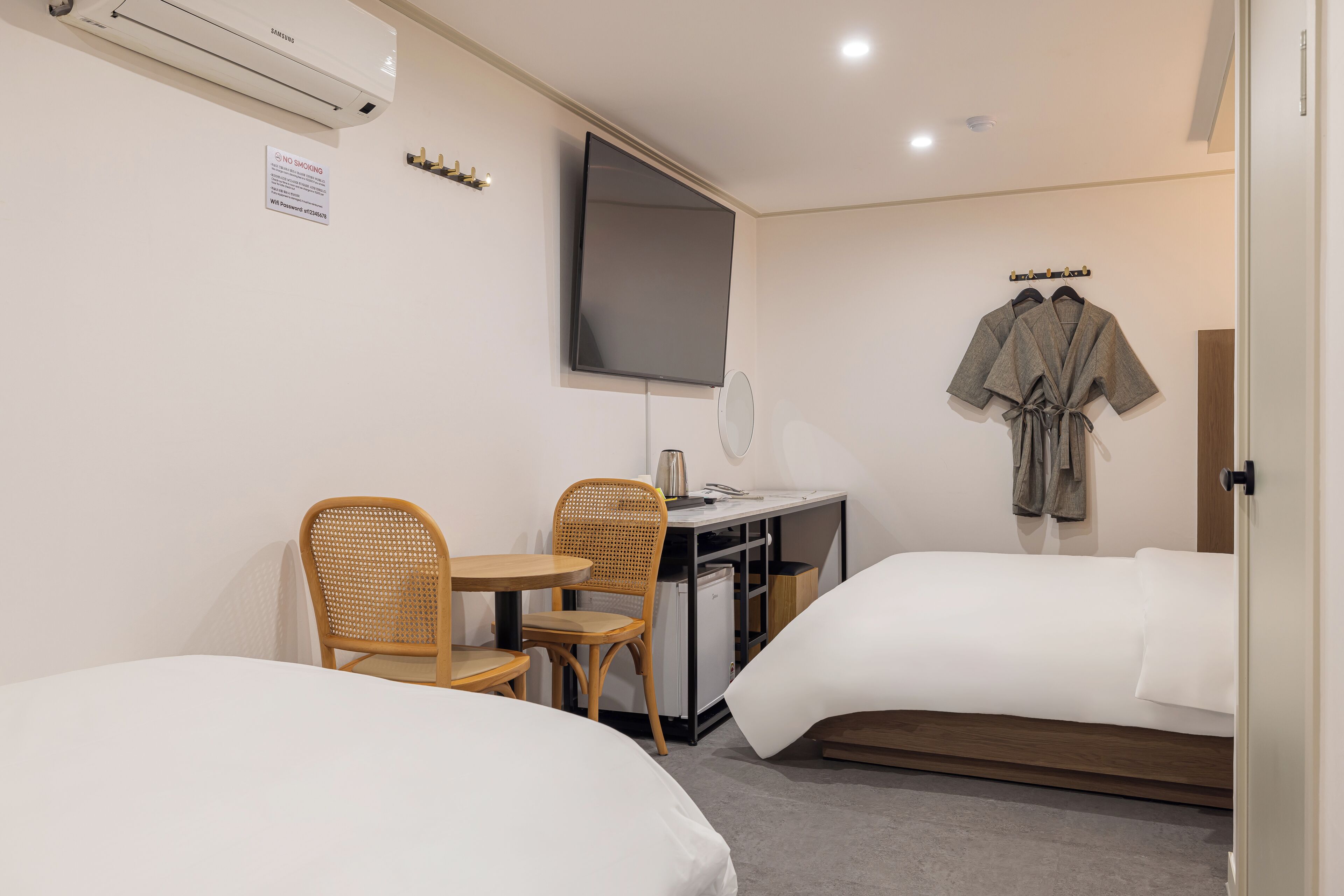 Habitación de hotel con dos camas, un escritorio con silla, un televisor montado en la pared y una bata colgada.