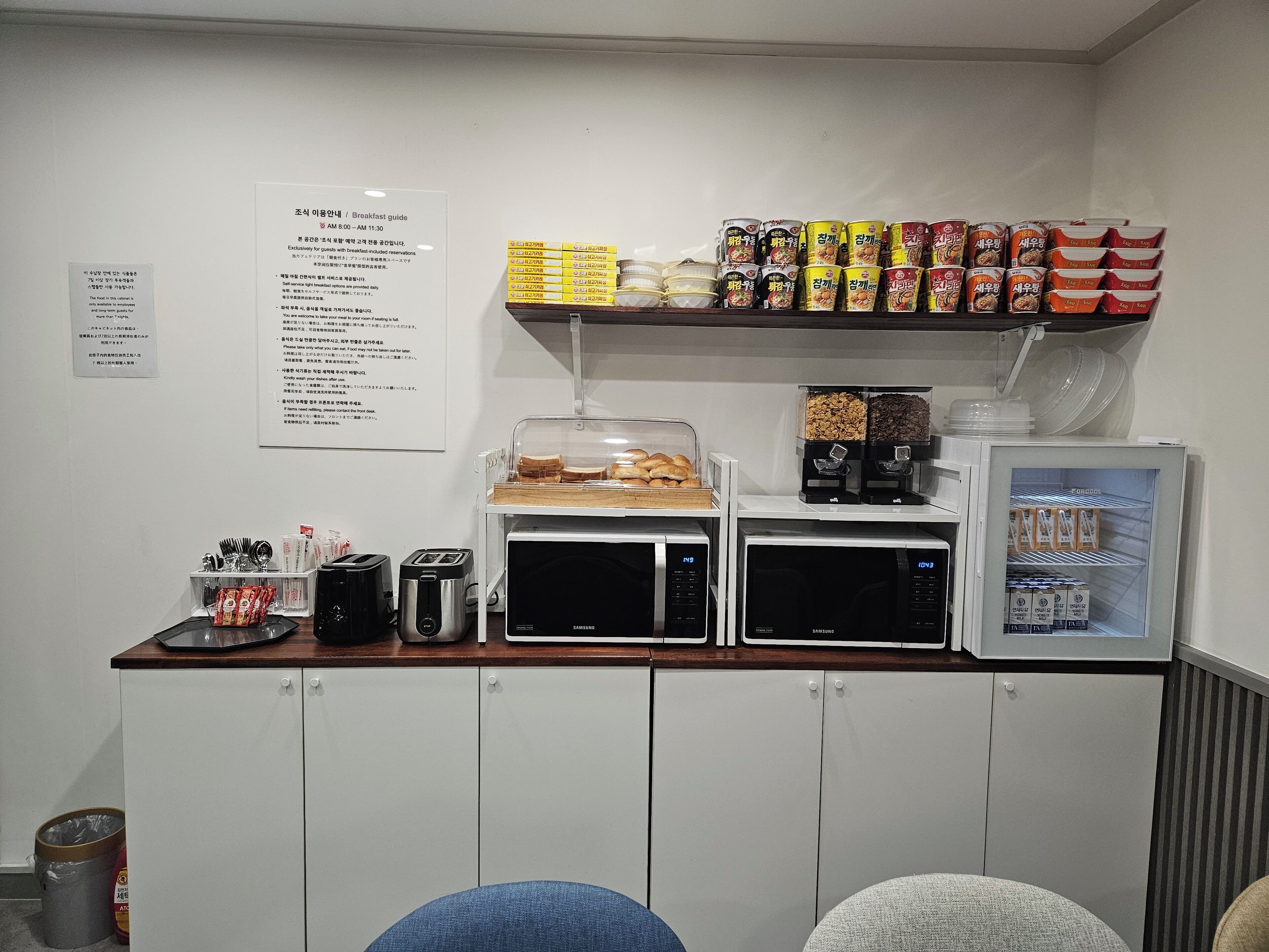 Un espace dédié au petit-déjeuner, équipé d’un four à micro-ondes, d’un grille-pain et d’une machine à café. On y trouve des étagères remplies de divers produits alimentaires et un réfrigérateur contenant des boissons.