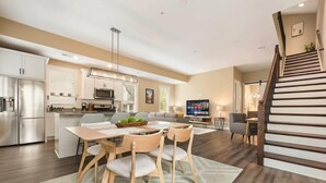 Eine moderne Küche mit Edelstahlgeräten, ein Essbereich mit Holztisch und -stühlen, ein Wohnzimmer mit Sofa und Fernseher sowie eine Treppe mit Holzstufen.