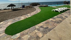 A patio with a stone and grass border, a pool, and lounge chairs facing the ocean. - Palacio Del Mar 102 - Rocky Point Oceanfront Vacation Rental (Puerto Peñasco)