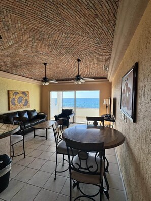 A room with a brick ceiling, a round dining table with chairs, a sofa, and a large window offering a view of the ocean. - Palacio Del Mar 207 - Rocky Point Oceanfront Vacation Rental (Puerto Peñasco)
