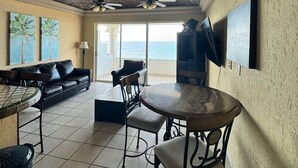 A spacious living area with a high ceiling, a round dining table, a black leather sofa, and a TV mounted on the wall. - Palacio Del Mar 307 - Rocky Point Oceanfront Vacation Rental (Puerto Peñasco)