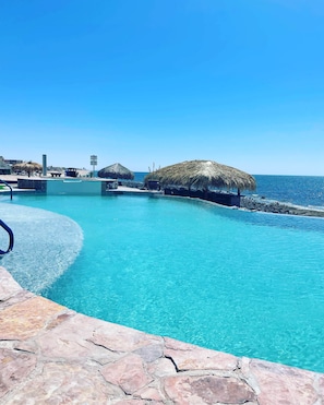 En klar, blå pool med stenkant, omgivet af stråtag parasoller og udsigt til havet.