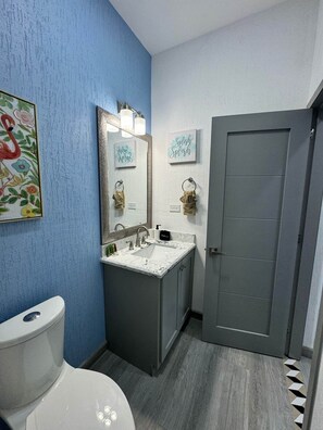 Ein Badezimmer mit blauer Wand, Toilette in Weiß, Waschbecken mit Spiegel und einer Tür.