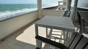 Ein überdachter Außenbereich mit Tisch und Stühlen mit Blick auf einen Strand und eine felsige Küstenlinie.
