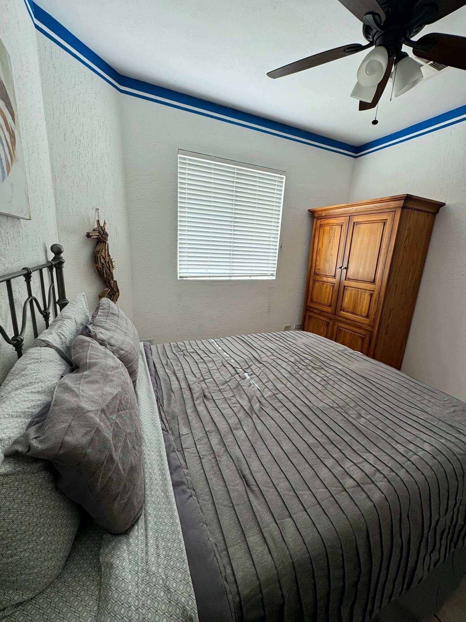 Une chambre à coucher avec un lit, un ventilateur de plafond, une fenêtre avec des stores et une armoire en bois.