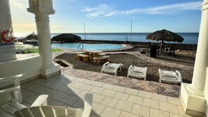 A covered outdoor seating area with white chairs and a table, overlooking a beach with a pool and umbrellas. - Palacio Del Mar 106 - Rocky Point Oceanfront Vacation Rental (Puerto Peñasco)