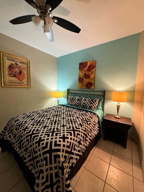 Une chambre à coucher comprenant un lit, deux lampes de chevet, un ventilateur de plafond, une affiche encadrée et un tableau floral accroché au mur.