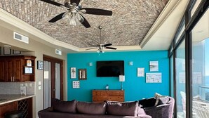 A modern living room with a ceiling fan, a sofa, a TV, and a view through a large window. - Palacio Del Mar 505- Rocky Point Oceanfront 4 Bedroom/4 Bath (Puerto Peñasco)