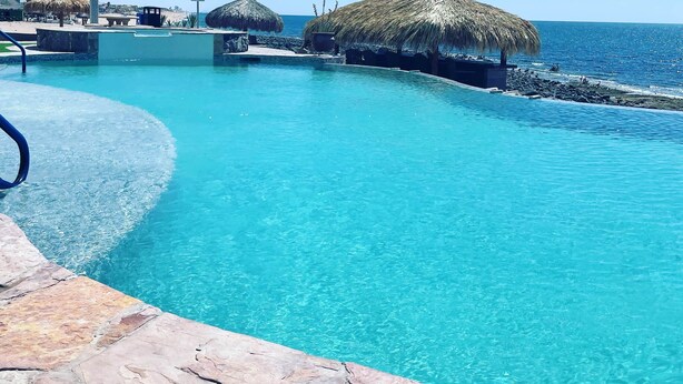 A clear blue pool with a stone edge, surrounded by thatched umbrellas and a view of the ocean. - Palacio Del Mar 505- Rocky Point Oceanfront 4 Bedroom/4 Bath (Puerto Peñasco)