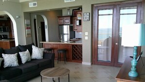 A living room with a sofa, a coffee table, and a kitchen area with a refrigerator and bar stools. - Palacio Del Mar 505- Rocky Point Oceanfront 4 Bedroom/4 Bath (Puerto Peñasco)