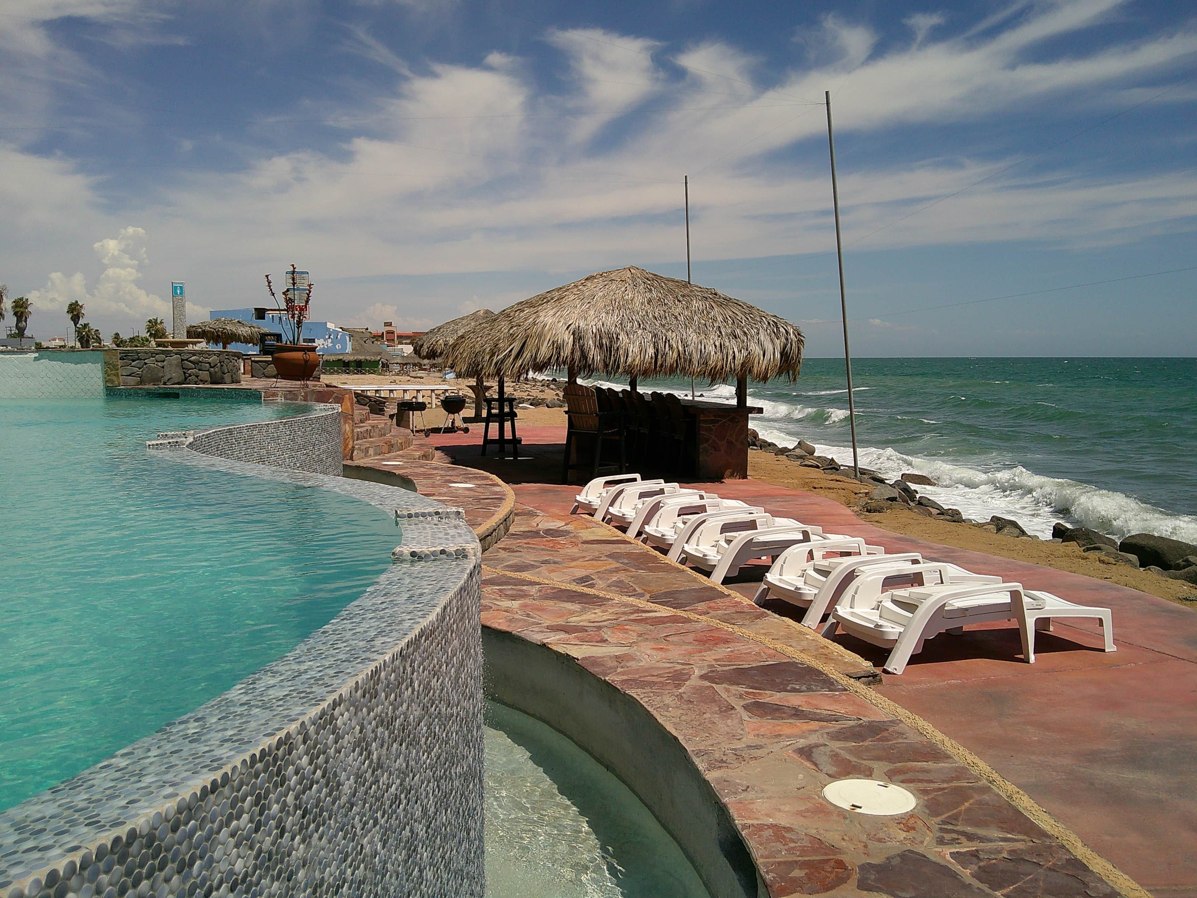 A pool area with a stone edge, a thatched umbrella, and white lounge chairs by the water.