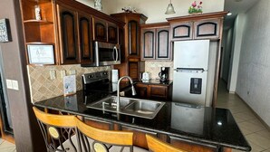 A kitchen with dark wooden cabinets, a black countertop, and a stainless steel sink. There are three bar stools with cushioned seats. - Palacio Del Mar 202 - Rocky Point Oceanfront Vacation Rental (Puerto Peñasco)