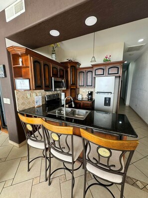 A kitchen with dark wooden cabinets, a black countertop, and a stainless steel sink. There are three bar stools with cushioned seats. - Palacio Del Mar 202 - Rocky Point Oceanfront Vacation Rental (Puerto Peñasco)