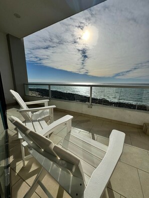 Un balcón con sillones blancos, barandilla de vidrio y vista al océano.