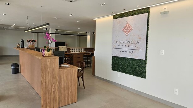 Zona de recepción de hotel con un mostrador de madera, un cartel en la pared que dice 'ESSENCIA HOTELS CAMPO ERÉ' y un área de estar con sillas y mesas.