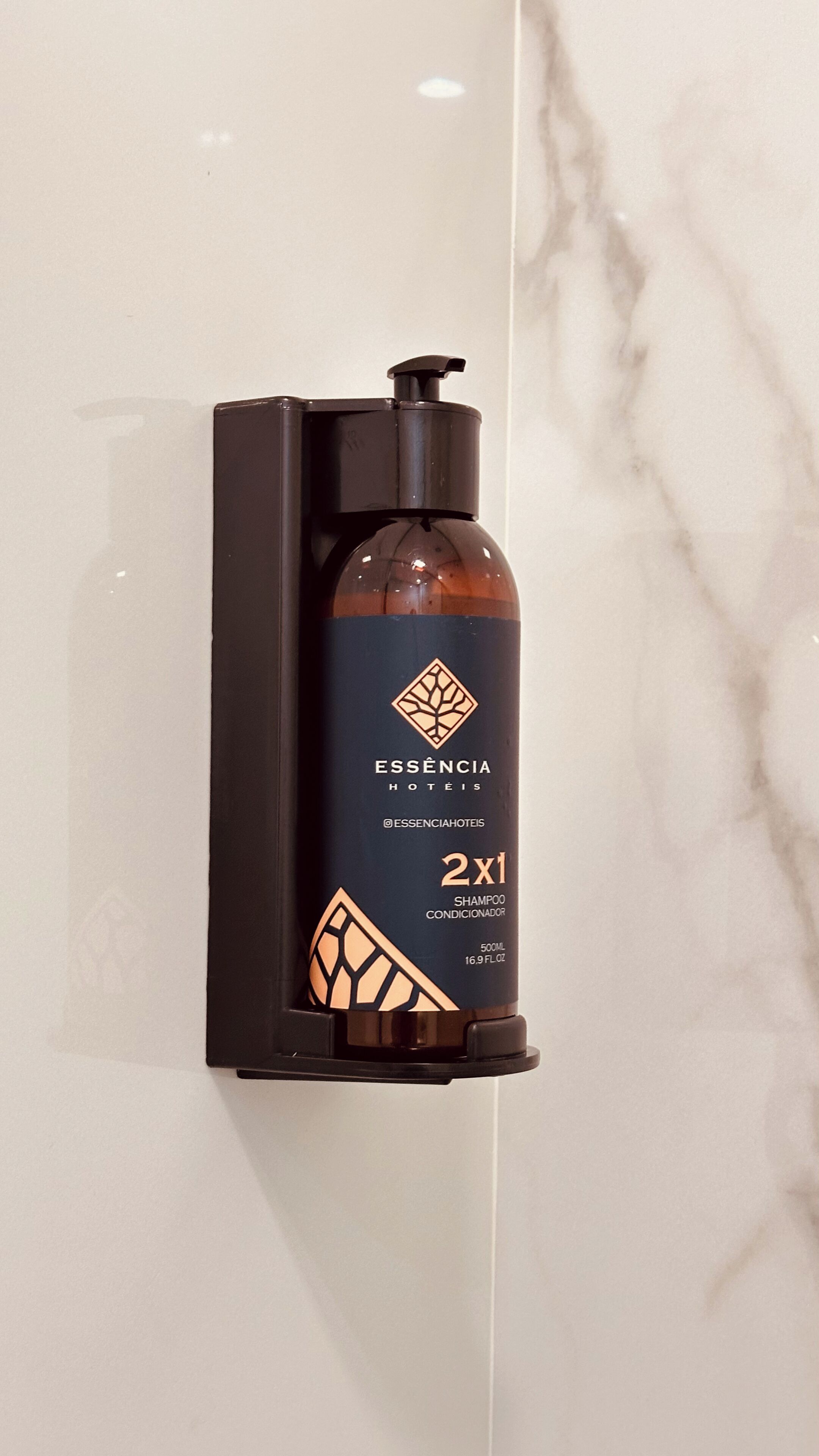 A black shampoo dispenser with a pump, labeled 'ESSENCIA HOTÉIS,' mounted on a white marble wall.