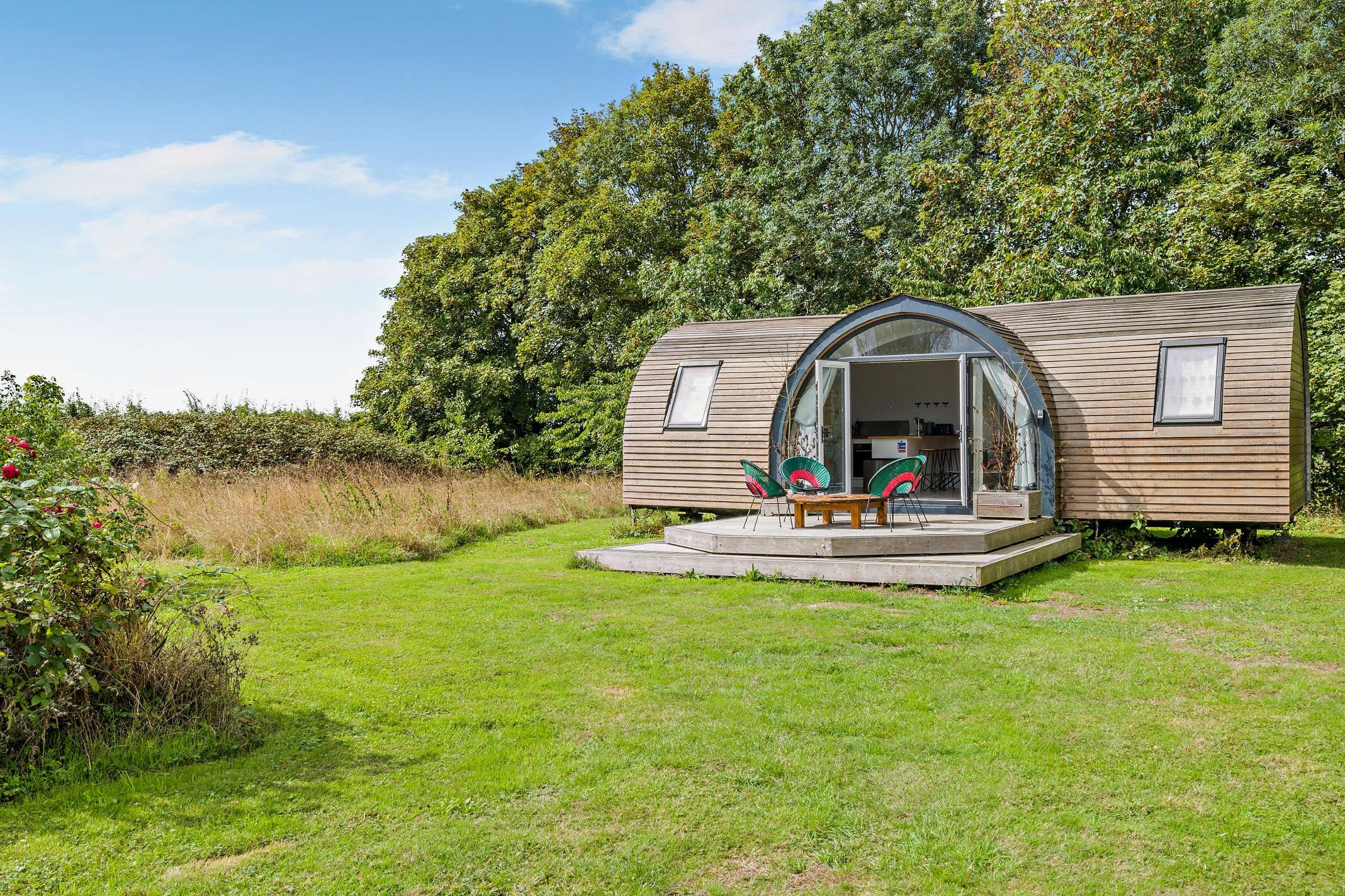 Une cabane en bois d’un seul étage, dotée d’une porte d’entrée en verre, d’un petit porche avec deux chaises et une table.