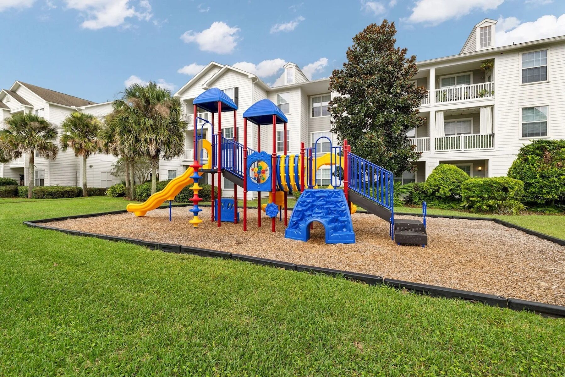 A playground with slides and climbing structures, surrounded by a grassy area and a building with balconies.