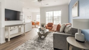 Uma sala de estar moderna com sofá, mesa de centro e televisão. Há uma área de jantar com cadeiras e mesa.
