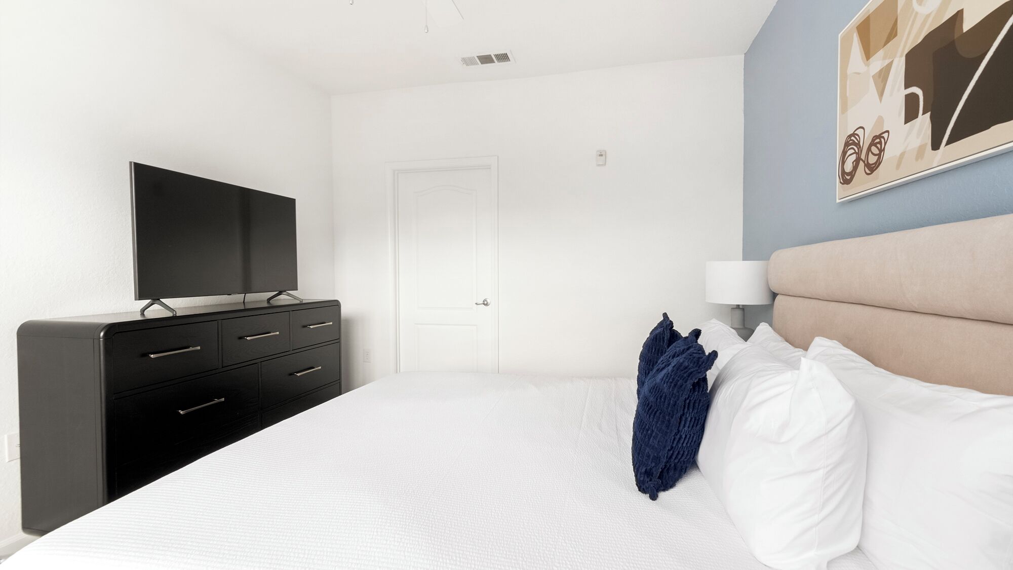 A hotel room with a bed, a TV on a dresser, a door, and a wall with a painting.