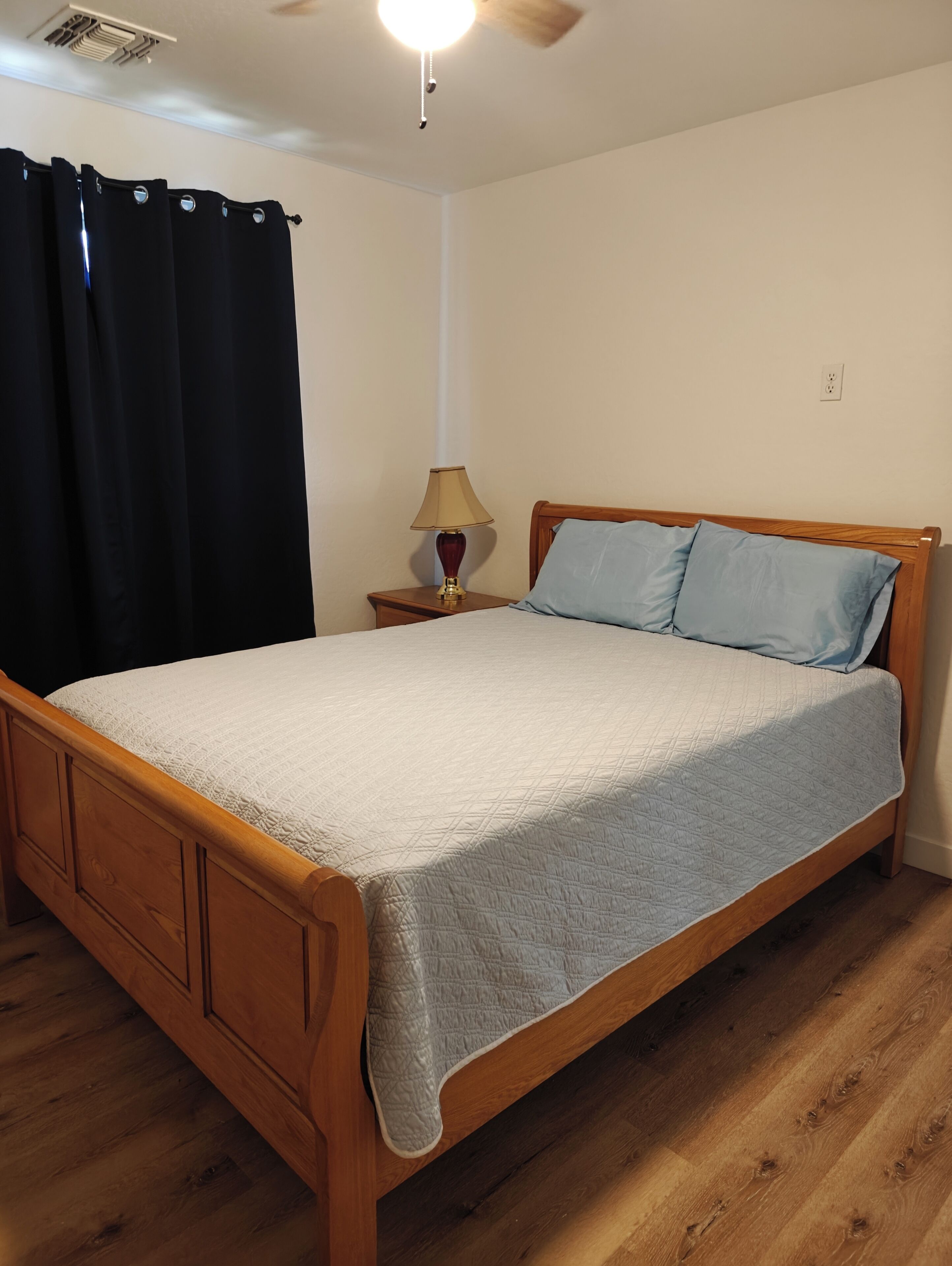 A bedroom with a wooden bed, a nightstand with a lamp, a ceiling fan, and a black curtain.