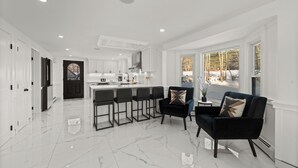 Una cocina moderna con armarios blancos, una isla central y taburetes de bar. Se observa una silla tapizada de negro con un cojín decorativo.