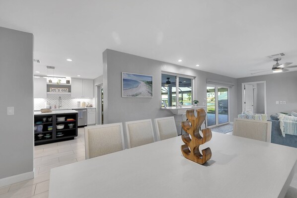A modern kitchen with white cabinets, a dining area with a white table and chairs, and a large window with a view of the outdoors. - 115 SW 57th Ter (Cape Coral)